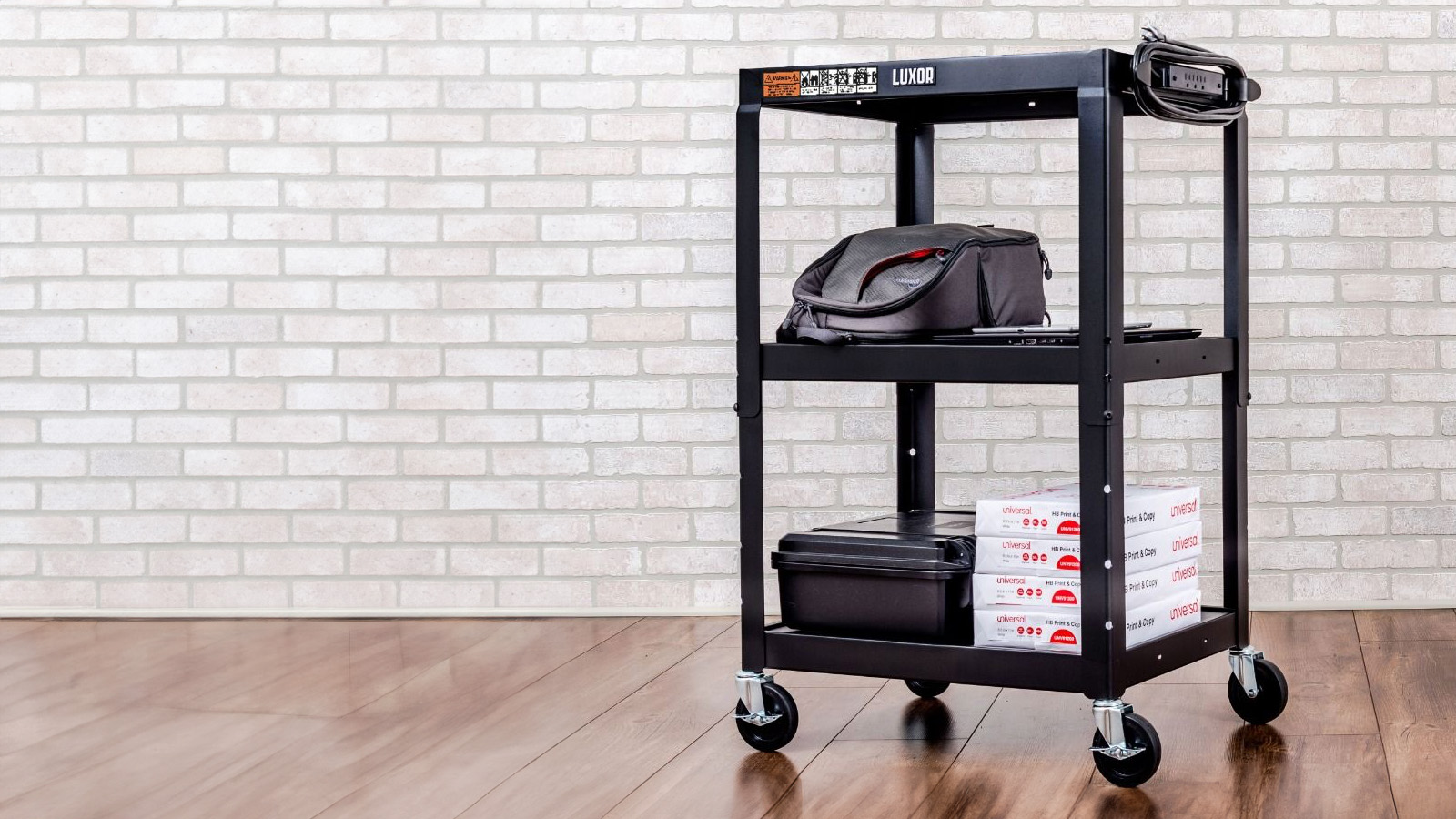 Luxor black metal multipurpose utility cart with wheels, featuring a power strip on top shelf, alongside office supplies and Universal print copy boxes on lower shelves, against a white brick wall.